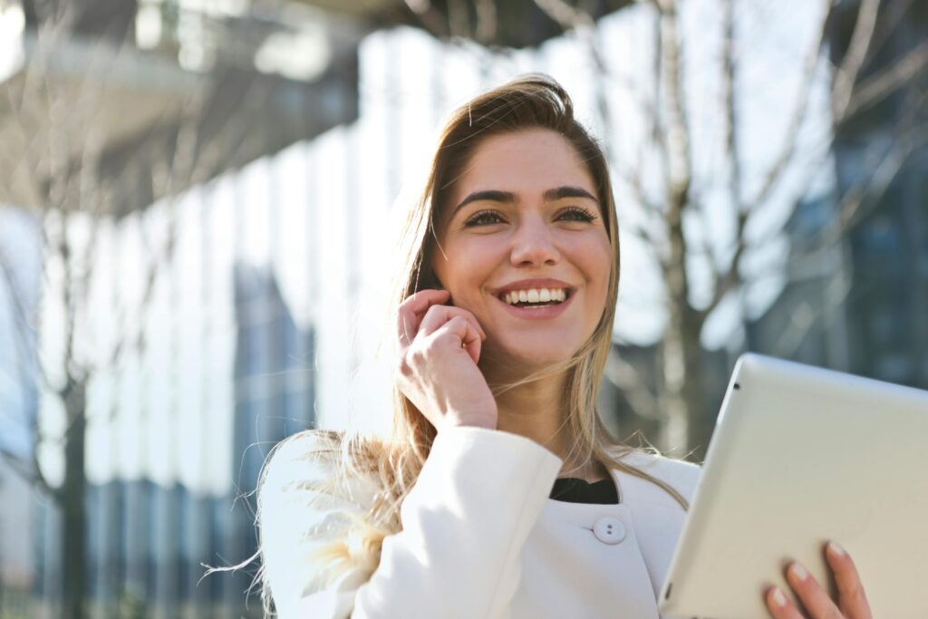 Confident businesswoman using her tablet and phone, smiling outdoors in sunlight. Contact us